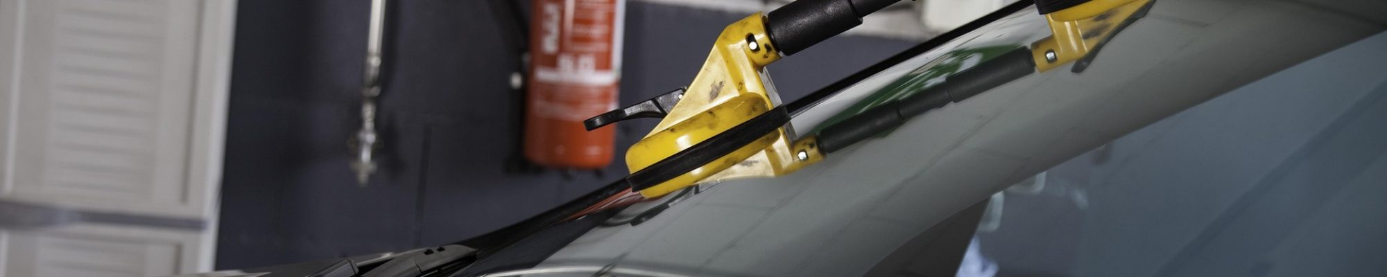 Twee gele zuignappen (glaslifters) worden gebruikt om een voorruit van een donkere auto te plaatsen tijdens het vervangen van de autoruit in de werkplaats van Autoschade Drenth in Amstelveen; op de achtergrond hangen een brandblusser en een EHBO-verbandtrommel aan de muur.